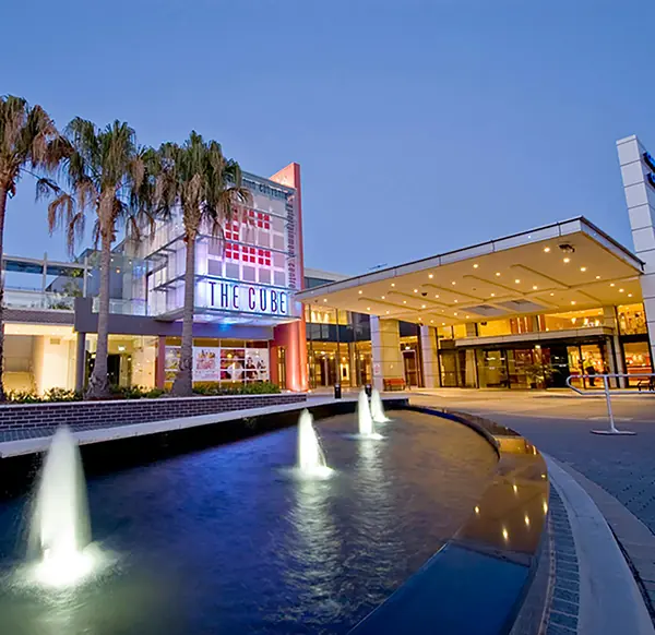 A modern building with “THE CUBE” sign, palm trees, fountains in the foreground, and a covered entrance, photographed at dusk—perfectly capturing the vibrant atmosphere surrounding Campbelltown land for sale and the latest land release in Cambelltown.
