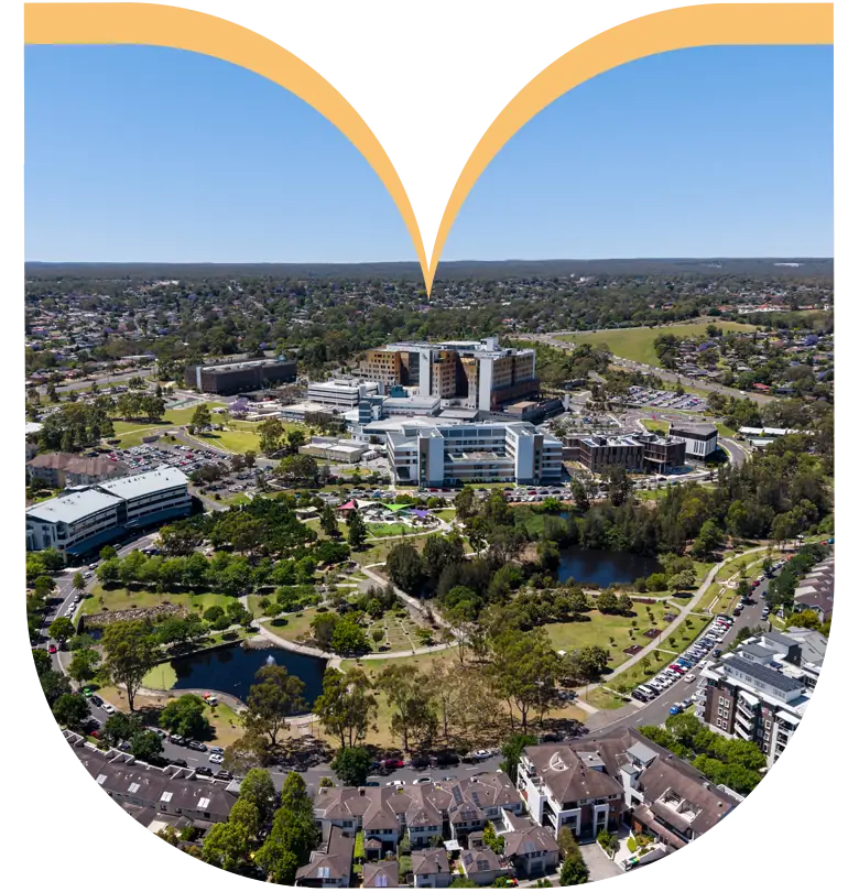 Aerial view of a large hospital complex surrounded by greenery, car parks, and residential buildings under a clear blue sky, with premium homesites featured in the adjacent new land development.