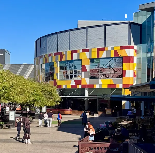 People walk near a modern building with a geometric yellow, red, and white façade under a clear blue sky. Trees and outdoor seating are visible in the foreground, adding to the vibrant atmosphere of this new land development.