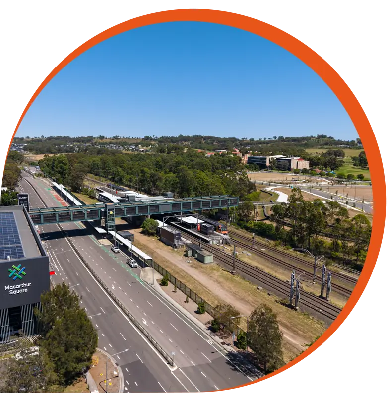 Aerial view of a railway station with trains on the tracks, adjacent roads, a footbridge, and surrounding greenery under a clear blue sky—an area poised for new land development and premium homesites.