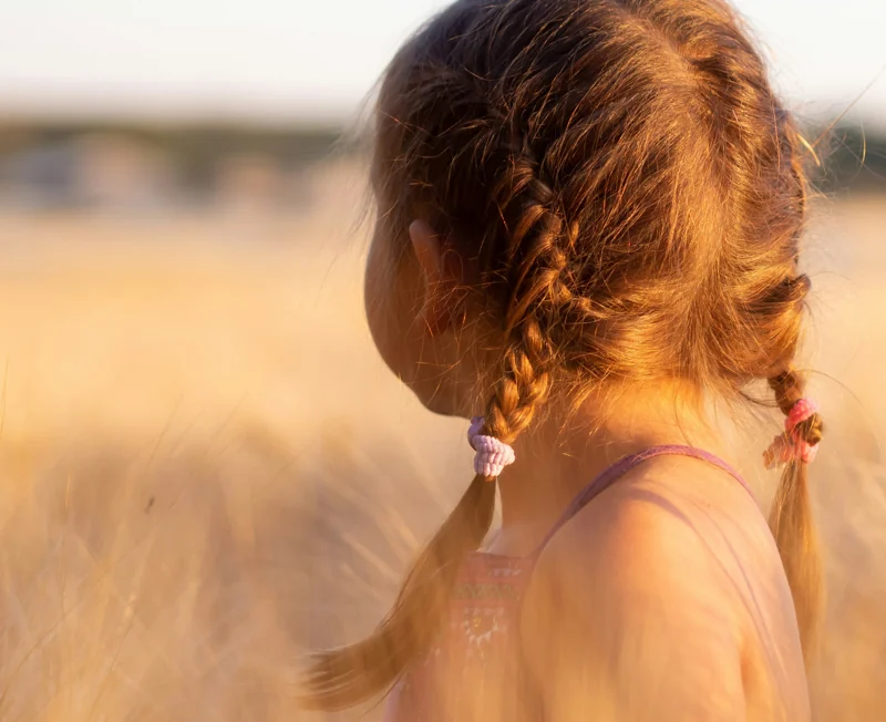 A young child with plaited hair stands in a sunlit field, facing away from the camera, where new Sydney homesites stretch beyond the horizon, evoking dreams of future possibilities.