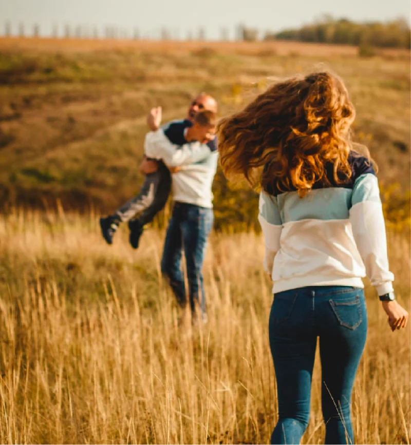 A woman walks towards a man spinning a child in tall dry grass, with fields and trees visible in the background, capturing the joy of family moments possible with the new land release in Campbelltown, where spacious landscapes invite new beginnings.